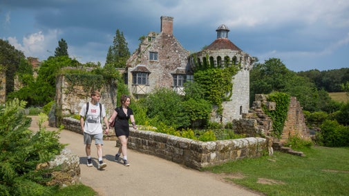 A young couple walking along a path in front of an old building with a two-storey round tower at one end and a gabled house at the other on a summer day